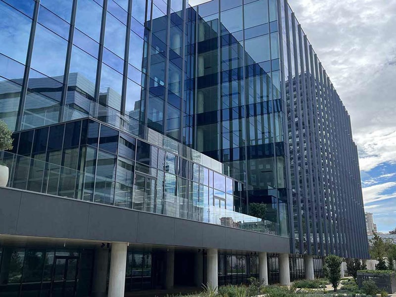 Modern glass buildings reflect the sky, surrounded by greenery, showcasing contemporary architecture in an urban landscape.